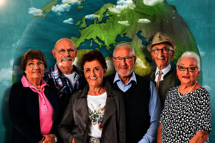 "Mit 80 Jahren um die Welt": Ruth, Nauke, Marianne, Theo, Ernst und Gisela stehen vor einer Weltkarte und lachen in die Kamera. Bild: Sender / ZDF / Svea Pietschmann / Alpenblick / KNSK. 